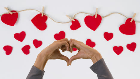 Valentine's-Day-Concept-With-Hands-Making-Heart-Shape-Against-White-Background-With-Red-Hearts-Attached-To-String